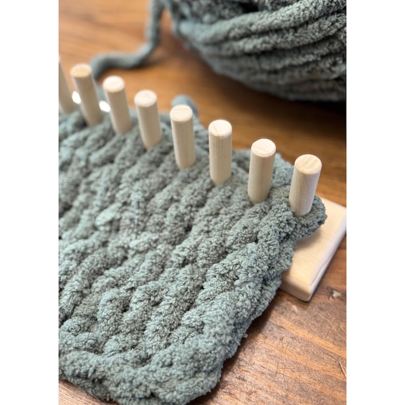 Op de afbeelding: Close-up van een saliegroen gebreid project op een houten breiraam. Het garen is dik en getextureerd, met houten pinnen die de steken op hun plaats houden. Een bol bijpassend garen is zichtbaar op de achtergrond, wat wijst op een knutselproject.