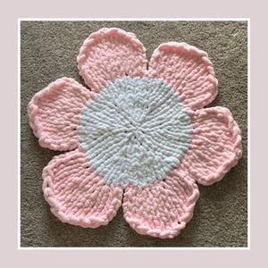 May include: A handmade, plush flower rug in a flower shape. The rug features a white center and pink petals. The petals have a textured, looped appearance. The rug is designed to be placed on the floor.