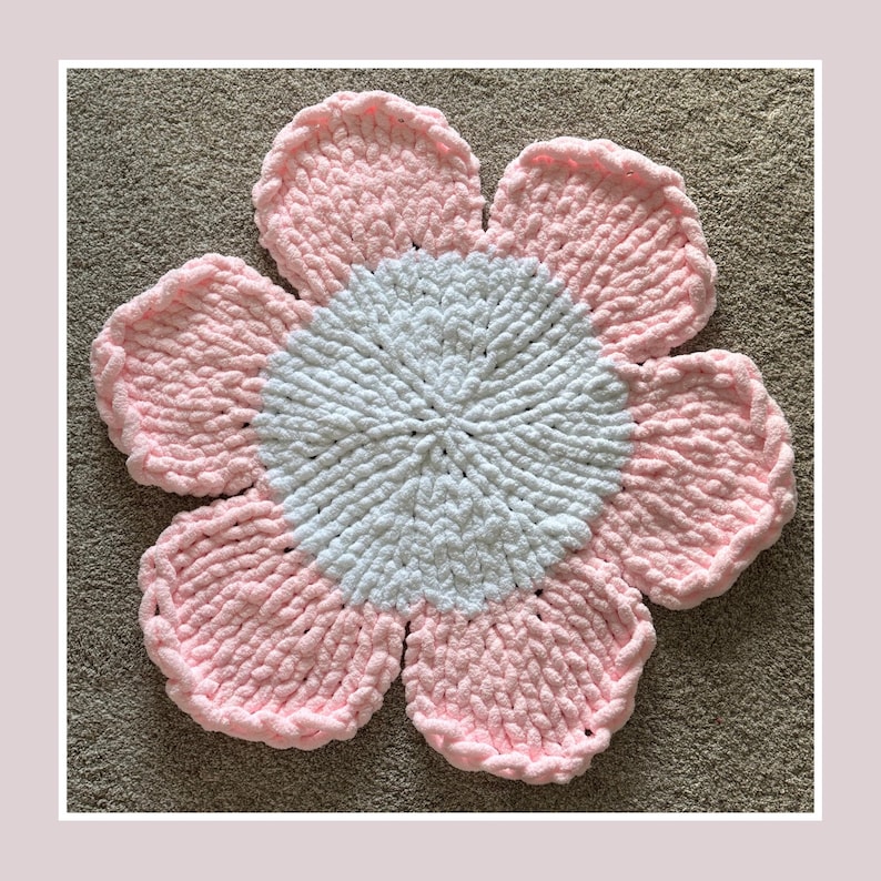 May include: A handmade, plush flower rug in a flower shape. The rug features a white center and pink petals. The petals have a textured, looped appearance. The rug is designed to be placed on the floor.