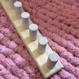 May include: A wooden peg rack with seven evenly spaced, cylindrical pegs. The rack is made of light-colored wood and is set against a textured, pink background. The pegs are also made of light-colored wood.