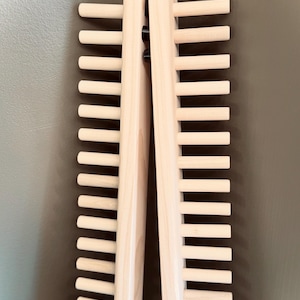 Op de afbeelding: Twee houten stropdashouders, elk met meerdere horizontale deuvels, worden getoond tegen een neutrale achtergrond. De houders zijn gemaakt van lichtgekleurd hout en zijn aan de bovenkant scharnierend. De deuvels zijn gelijkmatig verdeeld.