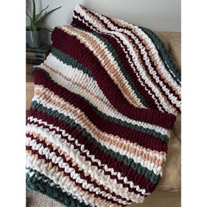 May include: A striped chunky knit blanket in shades of white, burgundy, green, and beige. The blanket is draped over a beige surface, highlighting the soft texture and colour variations. This cosy throw blanket is ideal for adding warmth and style to any living space.