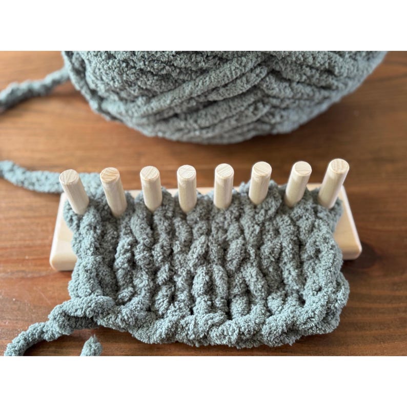 Op de afbeelding: Een close-up van een houten breiraam met een gedeelte gebreide stof in een zachte, gedempte groene kleur. Een grote bol bijpassend garen staat op de achtergrond. Het weefgetouw heeft houten pinnen en een lichtgekleurde houten basis.