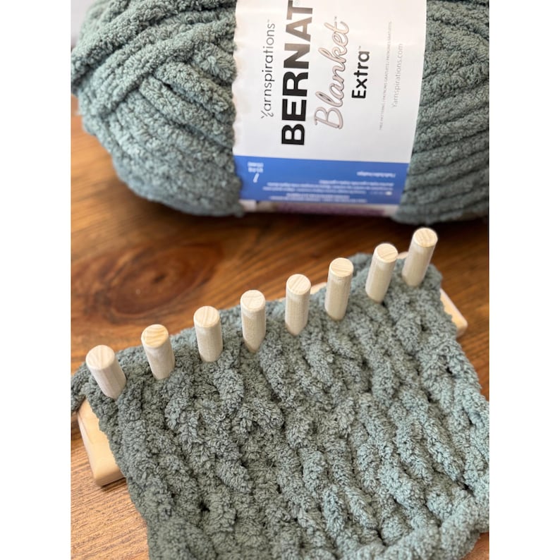 Op de afbeelding: Een bol Bernat Blanket Extra garen in een zachte groene kleur ligt achter een kleine houten breimachine met een gedeeltelijk voltooid project. Het garenlabel is zichtbaar. De weefgetouw heeft houten pinnen.