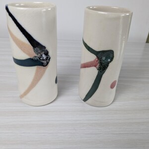 May include: Two off-white ceramic tumblers with abstract designs. One has black, navy, and tan brushstrokes, the other has green and red brushstrokes. Both have a small dot of color on the side.