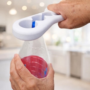 Könnte beinhalten: Ein weißer Flaschenöffner mit blauem Einsatz wird verwendet, um eine transparente Plastikflasche mit roter Flüssigkeit zu öffnen. Der Öffner wird von einer Hand gehalten.