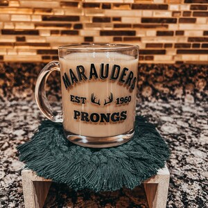May include: A clear glass mug filled with a light-colored beverage, featuring the text "Marauders Est 1960 Prongs" in a vintage style. The mug sits on a dark green macrame coaster atop a small wooden stand.
