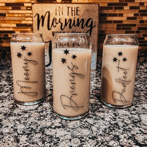 May include: Three clear glass beverage cups filled with a light brown liquid, each with a different name in a cursive font: "Moony", "Brongs", and "Badfoot". Each cup has a cluster of black star decals. A metal sign with the words "In the Morning" is in the background.