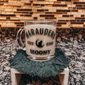 Könnte beinhalten: Ein klares Glas mit einem cremigen Getränk, mit dem Text "Marauders Est 1960 Moony" in Schwarz. Der Becher steht auf einem dunkelblauen Makramee-Untersetzer auf einem kleinen Holzständer, vor einem gefliesten Hintergrund.