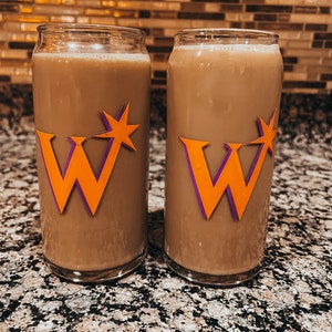 Puede incluir: Dos vasos de vidrio transparente llenos de un líquido marrón claro. Cada vaso presenta una gran "W" naranja con un contorno púrpura y un diseño de estrella. Los vasos están sobre una encimera moteada.