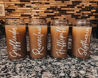 May include: Four clear glass tumblers filled with a light brown liquid. Each tumbler has a white cursive label with a different name: Gryffindor, Ravenclaw, Hufflepuff, and Slytherin. The tumblers are on a speckled countertop.