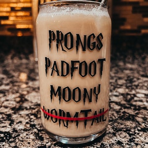 May include: A clear glass with a beverage and a metal straw. The glass has black text that reads "Prongs, Padfoot, Moony, Wormtail" with a red stripe. The glass is on a speckled surface.