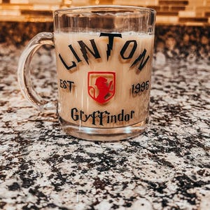 May include: A clear glass mug filled with a light brown beverage. The front features black text reading LINTON, a red shield emblem with a lion silhouette, the text EST 1995, and the word Gryffindor in black lettering at the bottom.