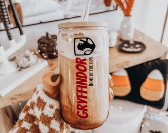 May include: A clear glass tumbler filled with a light brown iced coffee, featuring a bamboo lid and a clear glass straw. The glass is printed with a black lion crest and the red text Gryffindor Home of the Lion in a vertical orientation.