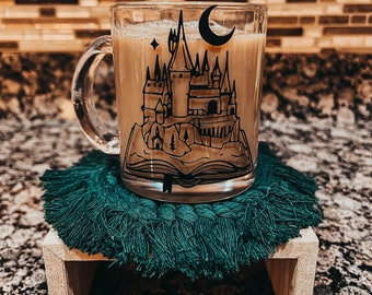 May include: A clear glass mug with a black castle design, filled with a light-colored beverage. The mug sits on a green macrame coaster atop a small wooden riser. The mug features a crescent moon and stars.