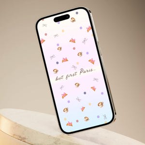 May include: A smartphone displays a pastel gradient wallpaper with scattered illustrations of coffee cups, bows, and pastries. The phrase "but first Paris..." is written in a cursive font. The phone is positioned on a white, angled stand against a neutral background.