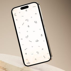 May include: A smartphone with a cream-colored screen displaying a repeating pattern of coffee cups, croissants, spoons, and hearts. The word "CAFE" is also scattered across the screen in a handwritten font. The phone is propped up on a white, rounded stand.