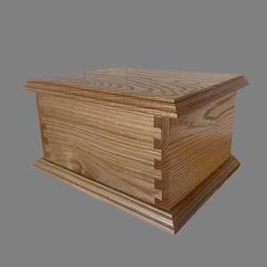 Op de afbeelding: Een houten doos met een natuurlijke houtkleur. De doos heeft een scharnierend deksel en een decoratieve rand rond de boven- en onderkant. De doos is gemaakt van eikenhout en heeft een natuurlijke houtnerf.