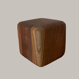 Könnte beinhalten: Ein massiver Holzquader mit abgerundeten Kanten. Die Holzmaserung ist sichtbar und zeigt eine Mischung aus hellen und dunklen Brauntönen. Das Objekt könnte ein dekoratives Element oder ein kleines Möbelstück sein.