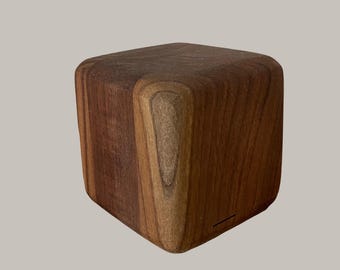 May include: A solid, wooden cube with rounded edges. The wood grain is visible, showcasing a mix of light and dark brown tones. The cube appears to be a decorative object or a small piece of furniture.