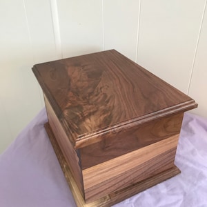 May include: A wooden box with a dark brown finish. The box is made of multiple pieces of wood, with a lighter brown wood used for the base. The box has a simple design with a raised edge around the top.