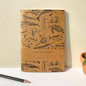 May include: A brown notebook with a sea life design. The cover features illustrations of fish, seahorses, crabs, and other marine creatures. The text on the cover reads "PLAIN NOTEBOOK 148gsm recycled paper" and "dopkit zivera designs".