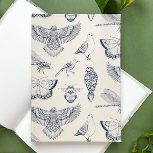 Op de afbeelding: Een crèmekleurig notitieboek met een blauw en wit patroon van vogels, vlinders en libellen. Het notitieboek heeft een gestippeld interieur.