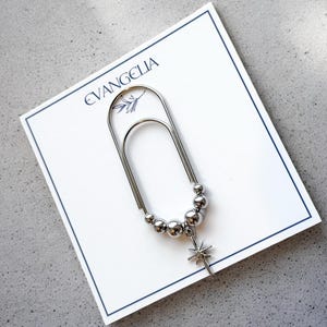 May include: A silver-toned paper clip with a beaded chain and a star charm, resting on a white card with the word "EVANGELIA" printed in blue. The card has a blue border and is set against a gray surface.