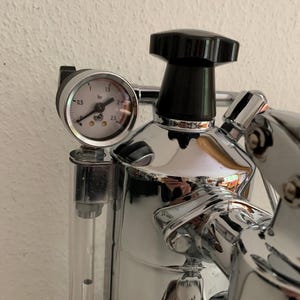 Könnte beinhalten: Nahaufnahme einer verchromten Espressomaschine mit schwarzem Knopf und Manometer. Das Manometer zeigt den Druck in bar an, von 0,5 bis 2,5. Ein durchsichtiges Rohr ist seitlich sichtbar.