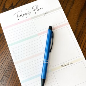 May include: A white notepad with the text "Today's Plan" at the top. The notepad is divided into three sections: Goals, To-Dos, and Reminders. A blue pen is lying on top of the notepad.
