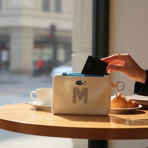 May include: A cream-colored pouch with a rhinestone "M" on a wooden table. A hand is reaching into the pouch, and a cup of coffee and a croissant are nearby. The scene is set in a cafe.