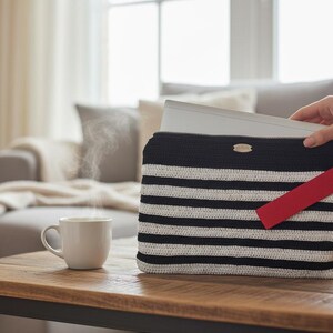 Puede incluir: Un bolso de crochet a rayas blancas y negras con una etiqueta roja, que contiene un ordenador portátil. Una taza blanca con vapor está sobre una mesa de madera. El bolso mide unos 30 cm de ancho.