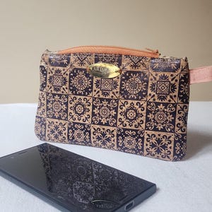 May include: A rectangular cork pouch with a repeating geometric pattern in dark blue and gold. The pouch has a peach-coloured zip and a small peach-coloured handle. A smartphone is in front of the pouch.