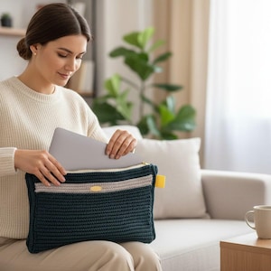 Puede incluir: Una funda para portátil de ganchillo azul oscuro con una raya beige y crema y una lengüeta amarilla. La funda la sostiene una persona que lleva un jersey crema. Se está colocando un portátil dentro de la funda.