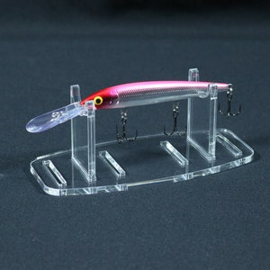 Puede incluir: Un soporte de exhibición de señuelos de pesca de acrílico transparente con un señuelo de pesca rosa y plateado con tres anzuelos.