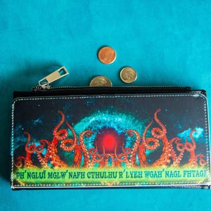 May include: Black wallet with a zipper, featuring a cosmic design with red tentacles and the text "PH'NGLUI MGLW' NAFH CTHULHU R'LYEH WGAH'NAGL FHTAGN". Several coins are placed on a teal surface.