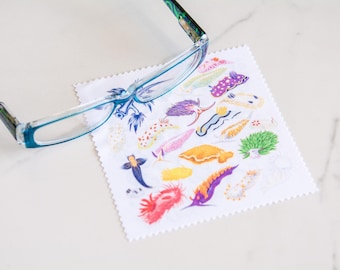 May include: A pair of eyeglasses rests on a white, patterned cleaning cloth. The cloth features colorful illustrations of sea slugs. The glasses have a blue frame and patterned temples. The cloth has a scalloped edge.