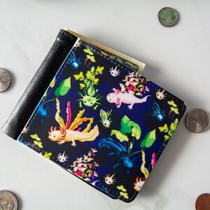 May include: A black bi-fold wallet with a colorful axolotl design. The wallet features a dark blue background with illustrations of axolotls, aquatic plants, and other aquatic life. Several coins are scattered around the wallet.