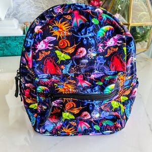 May include: A brightly patterned backpack with a marine life design. The bag showcases a variety of sea creatures, including axolotls, fish, and snails, in a mix of red, blue, yellow, and green on a black backdrop. It has a front zip pocket and black zips.