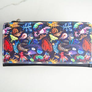 May include: A rectangular wallet with a black base and a zipper closure. The wallet's surface is covered with a colorful pattern of various sea creatures, including axolotls, snails, and fish, set against a dark blue background.