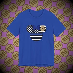 以下が含まれることがあります： ロイヤルブルーのTシャツに、ブルーラインとアメリカ国旗のミッキーマウスのデザインが施されています。ミッキーマウスの耳は、アメリカ国旗とブルーラインの旗でいっぱいです。リボンは紫です。