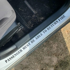 May include: A white car door sill with the text "PASSENGER MUST BE ABLE TO RETURN FIRE" in black. The car's interior includes a gray seat and a black floor mat with "DODGE" printed on it. The car door is open, revealing grass.