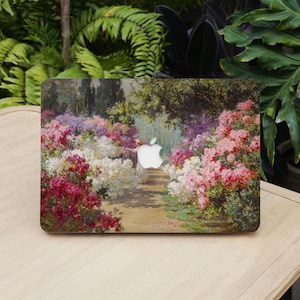 May include: A laptop cover featuring a vibrant painting of a garden path lined with blooming flowers in shades of red, pink, white, and purple. The Apple logo is visible. The cover is on a light-colored wooden table, with green foliage in the background.
