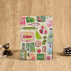 May include: A decorative case featuring a watercolor illustration of various food items, including sardines, figs, olives, and a bottle of sparkling water. The case has a beige background and is surrounded by small figurines and a pinecone.