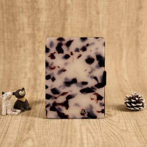 May include: A rectangular e-reader case featuring a white and translucent base with a dark brown and black tortoiseshell pattern. The name ELEANORA is printed in small gold capital letters near the bottom center of the front cover surface.
