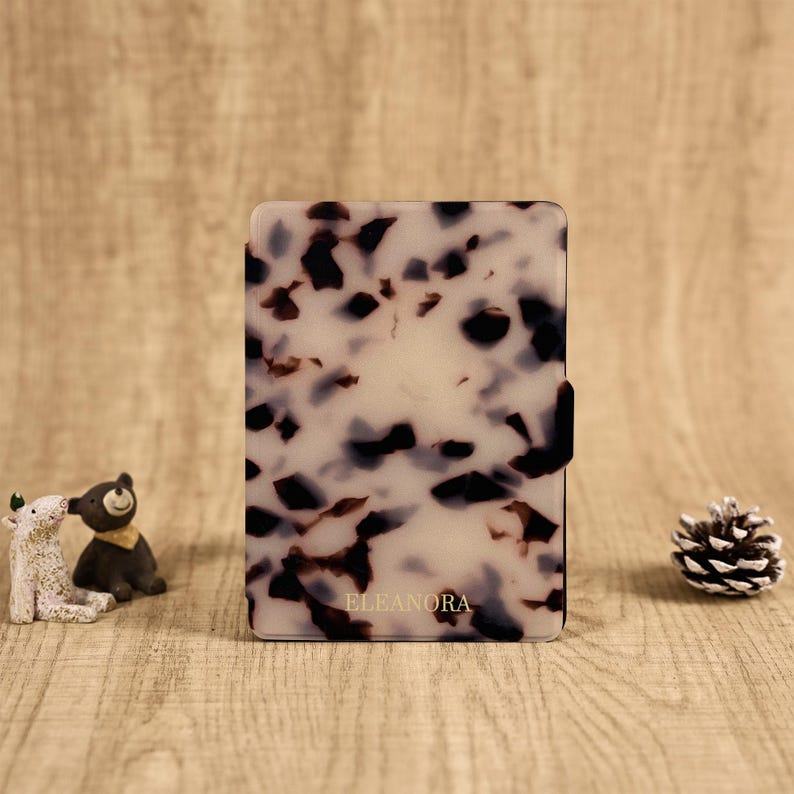 May include: A rectangular e-reader case featuring a white and translucent base with a dark brown and black tortoiseshell pattern. The name ELEANORA is printed in small gold capital letters near the bottom center of the front cover surface.