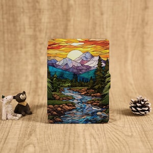 May include: A colorful wallet featuring a stained-glass design of a mountain landscape with a river, trees, and a sunset. The color palette includes shades of blue, purple, orange, and green. The wallet is rectangular and has a dark border.