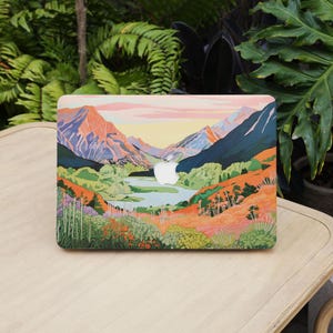 May include: A laptop skin featuring a colourful illustration of a mountain range with a lake in the foreground. The mountains are a mix of blue, green, and orange, and the lake is a light blue. The sky is a light pink and orange.