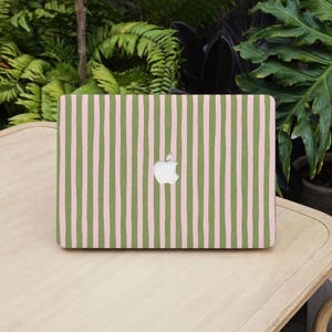 May include: A laptop with a green and pink striped pattern. The Apple logo is visible in the center. The laptop is on a light-colored wooden surface, with green foliage in the background.