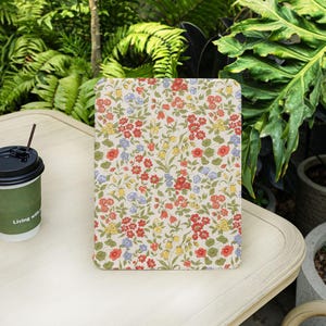 May include: A floral patterned tablet case featuring red, blue, and yellow flowers on a cream background. The case is rectangular with rounded corners and sits on a white table. A coffee cup with a straw is to the left.